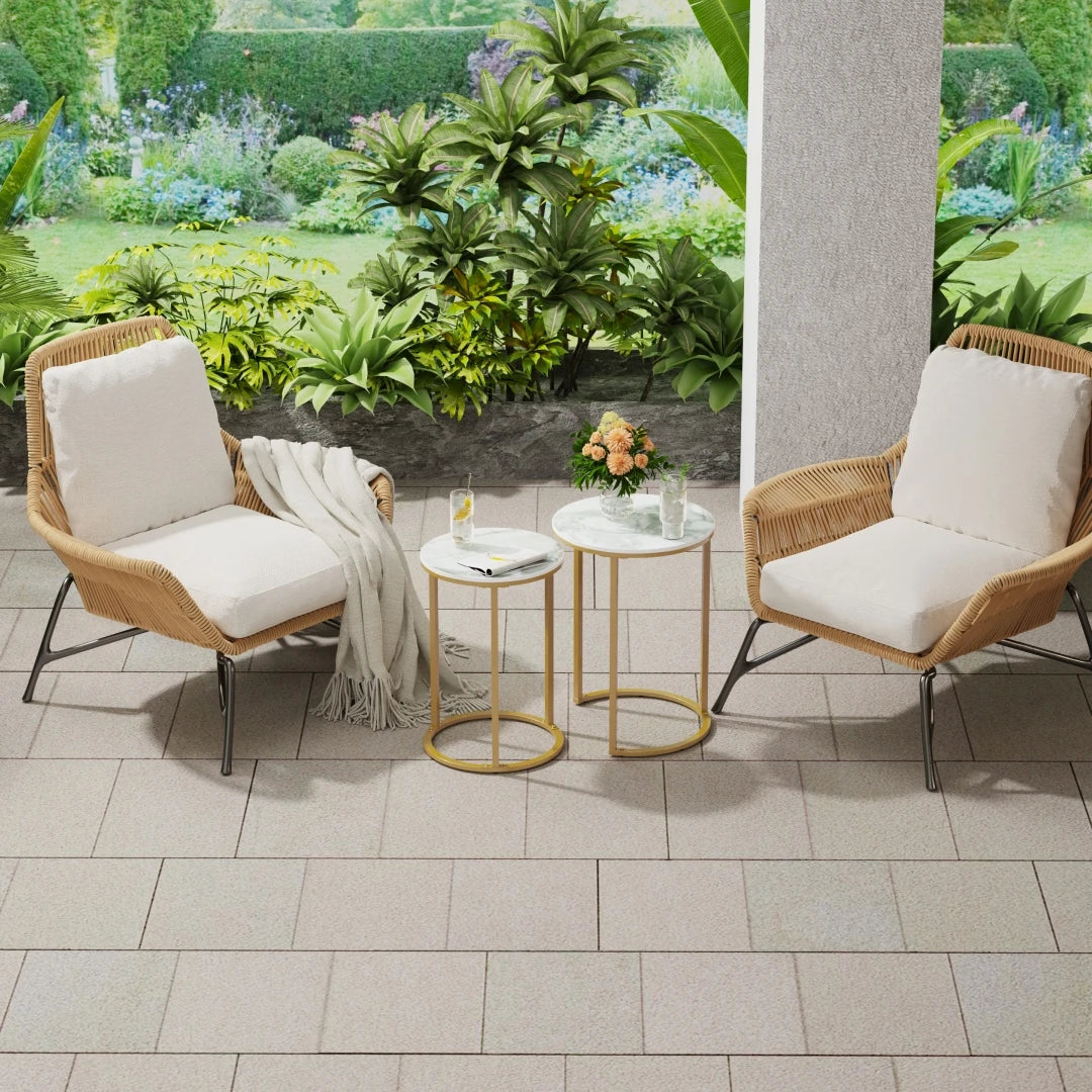 Two wicker chairs with white cushions and small tables on a tiled patio with garden view.